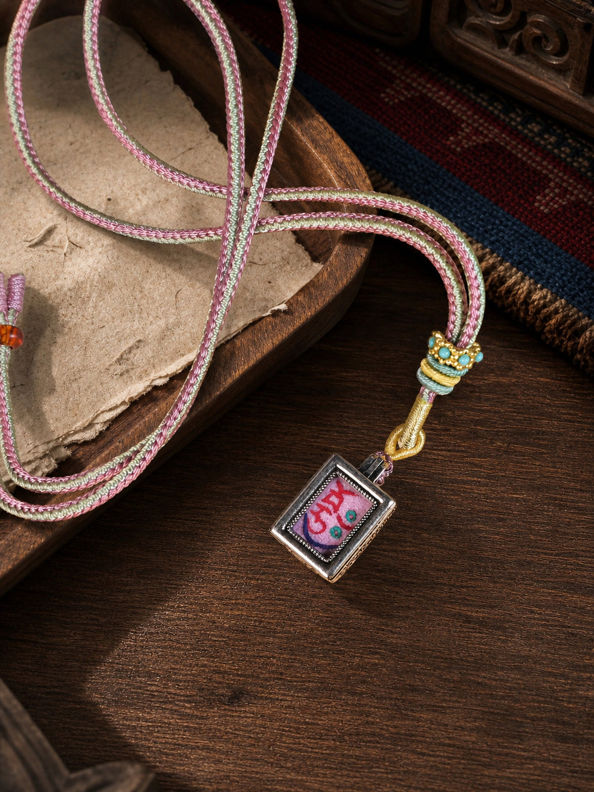 Zaki Lhamo — Style A design mini thangka pendant: painted symbol on pink, with a pink-and-cream braided cord