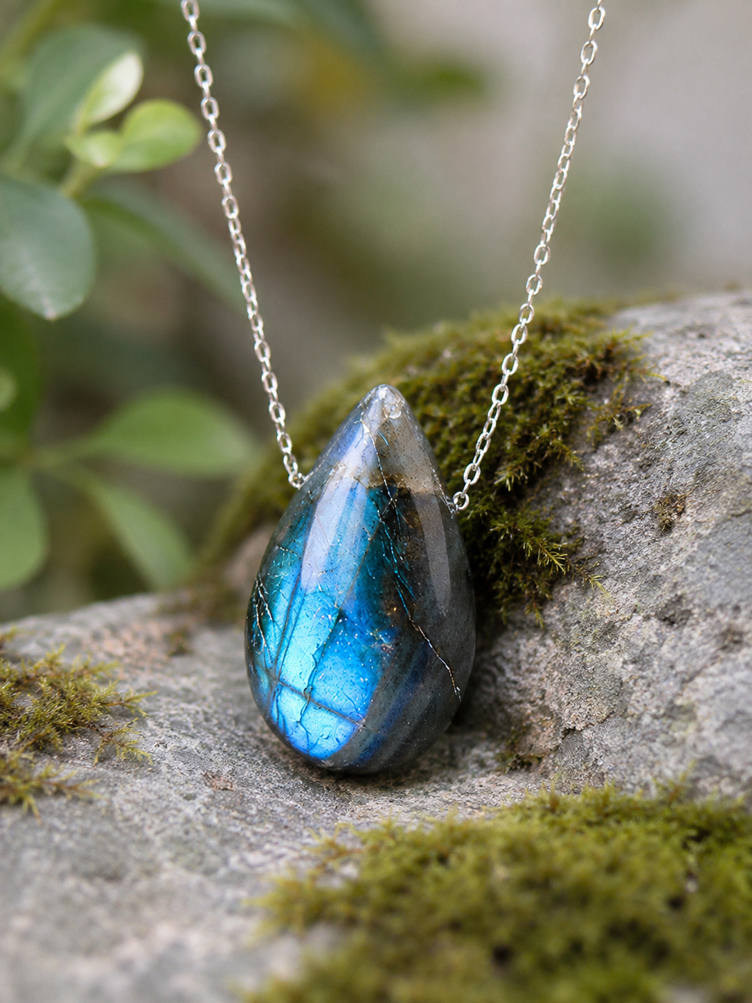 Blue-flash teardrop pendant shown on a silver-colored chain against mossy rock