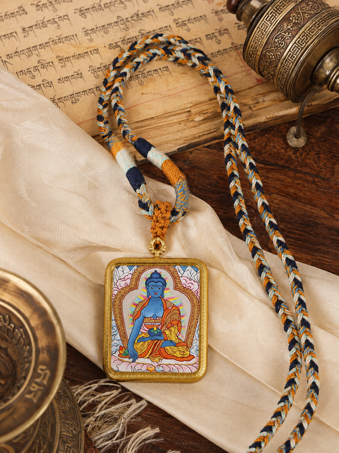 Medicine Buddha pendant and braided cord arranged on cream fabric beside printed pages